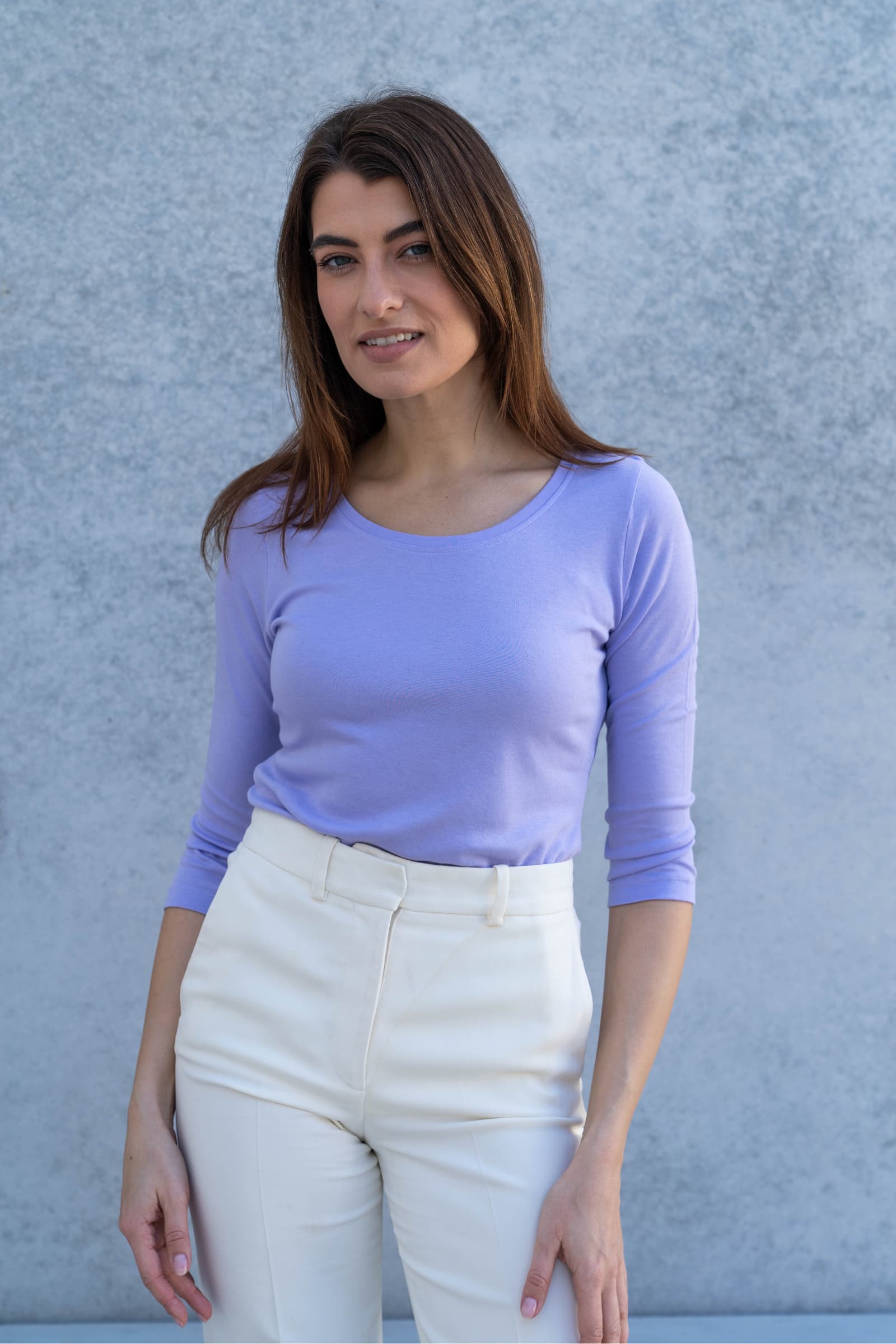 Woman wearing a light purple long-sleeve top and white pants against a gray background