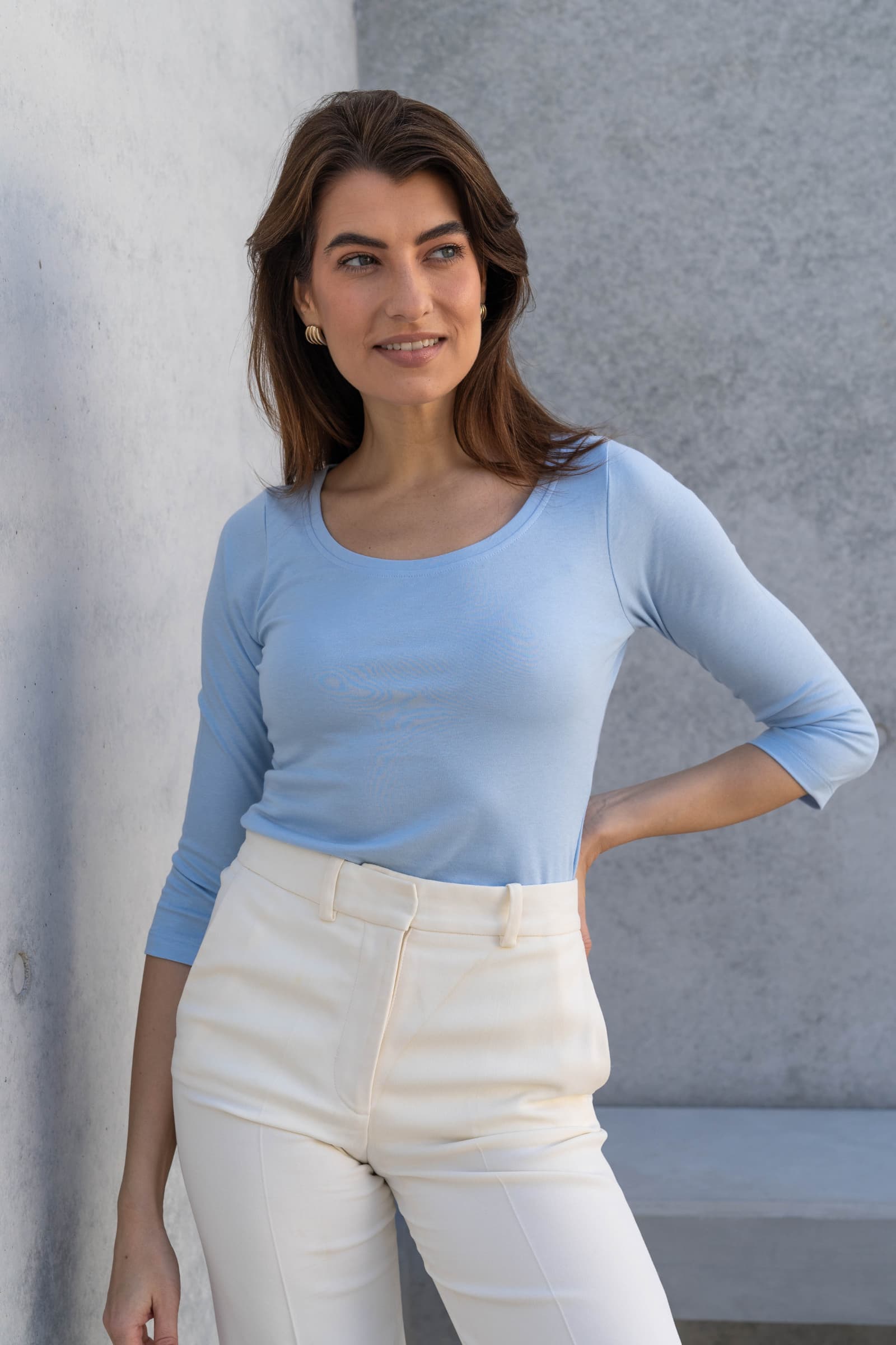 Woman wearing a light blue top and white pants against a gray wall.
