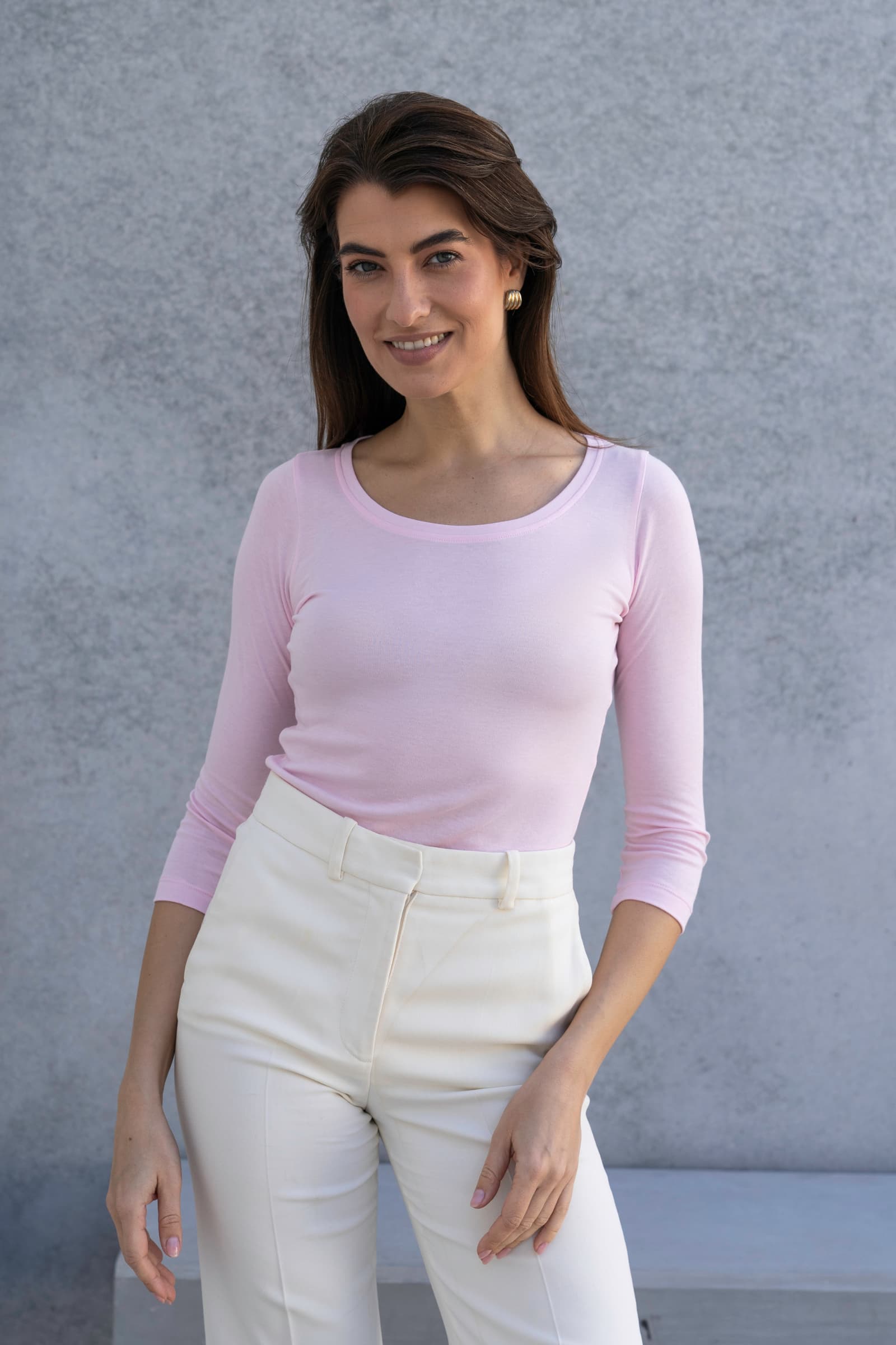 Woman wearing a light pink long-sleeve top and white pants against a gray background